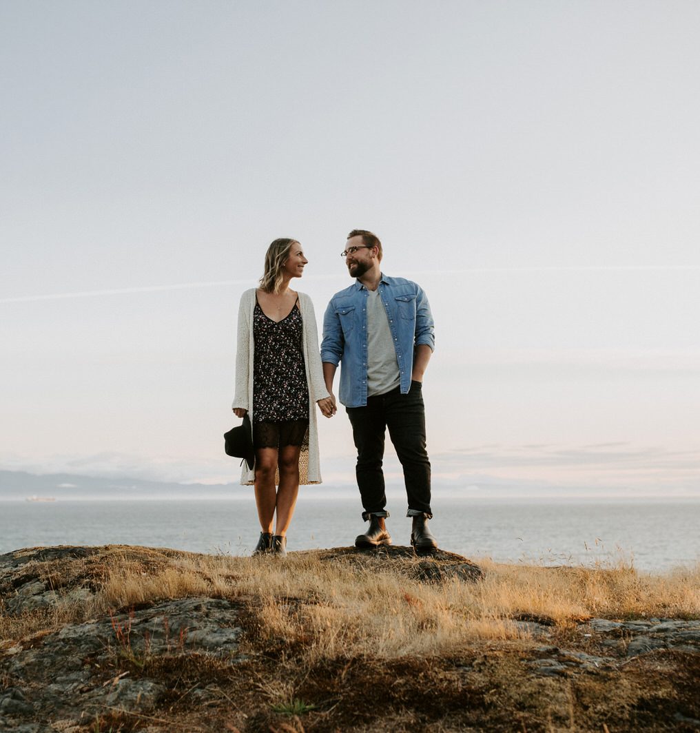 couple stands on a rock in the PNW overlooking the ocean at sunset for their engagement photos
